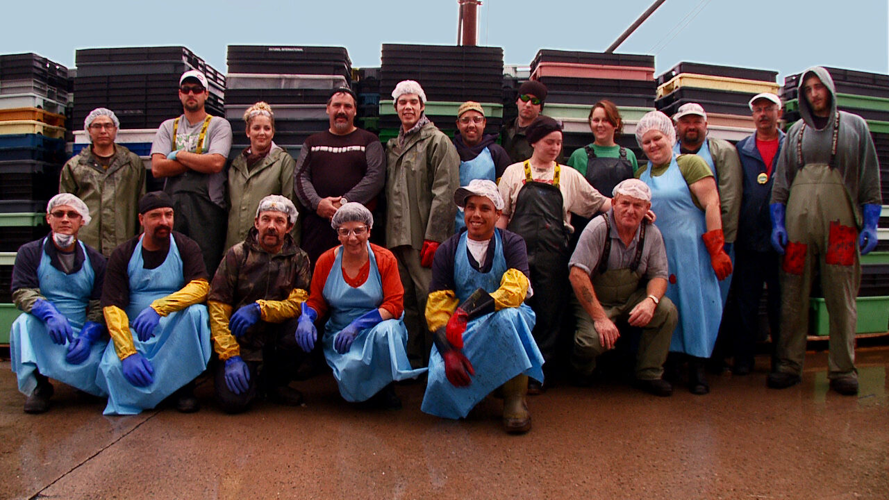 group of fish processing plant workers pose outside in their work aprons and gloves in front of fish crates