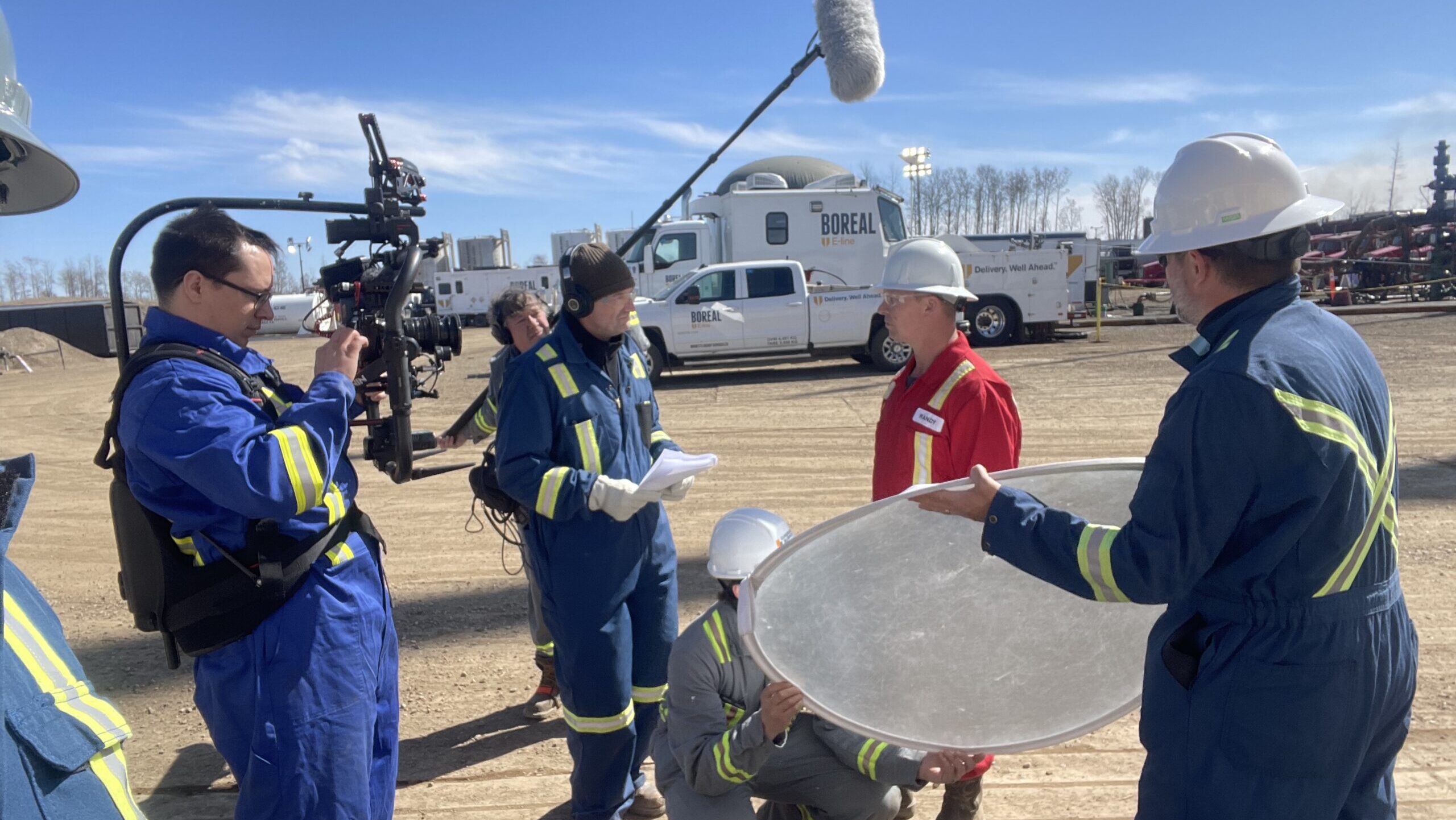 film crew with camera on steady cam apparatus, with sound recordist microphone, assistant with reflector, all in safety gear, filming a worker on location