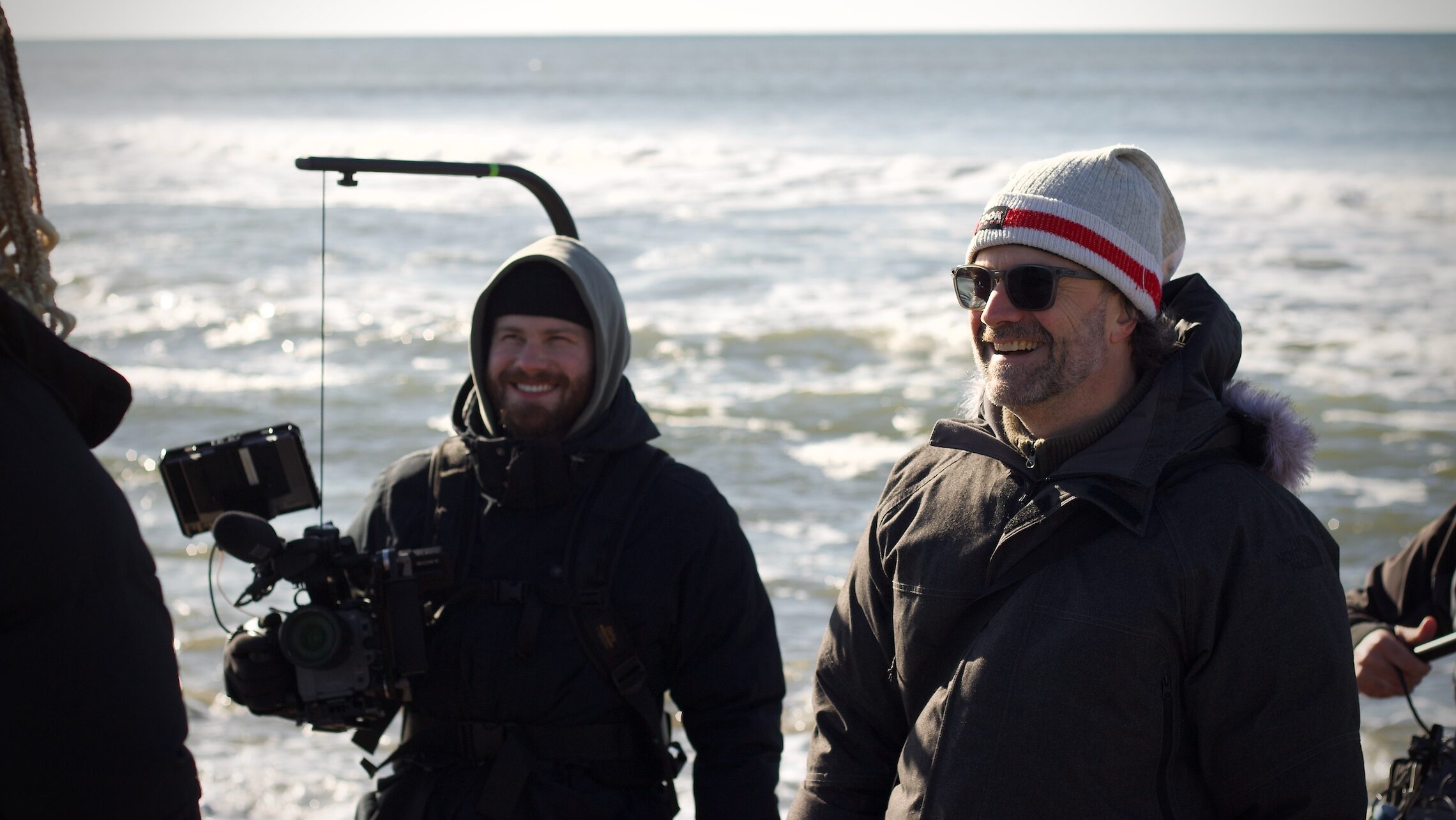 director and cameraman stand together laughing with on camera participants on beach, ocean in background